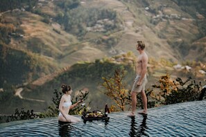 A couple by an infinity pool with a scenic view of hills and a tray with food and drinks.