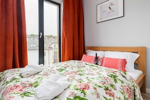 Ein Hotelzimmer mit Bett, Blumenbettwäsche, einem Holzkopfstück, einem Fenster mit Vorhängen und einem gerahmten Bild an der Wand.