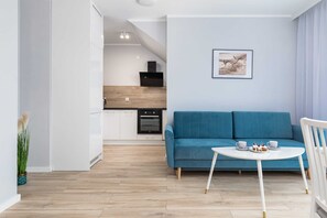 Ein modernes Wohnzimmer mit einem blauen Sofa, einem kleinen runden Tisch mit Tassen und einem Teller sowie einer Küche im Hintergrund.
