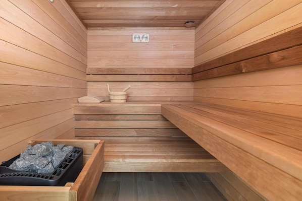 A wooden sauna with benches, a stone basin, and a black container with stones.