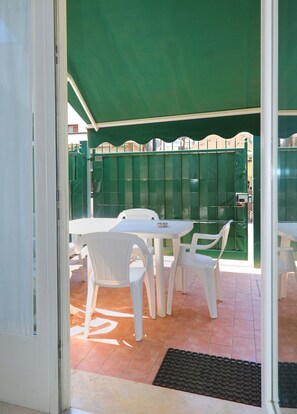 A patio area with a white table and chairs, a green awning, and a view of a building in the background.