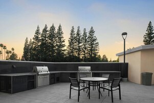 Un patio exterior con una parrilla, asientos y vistas a árboles altos.