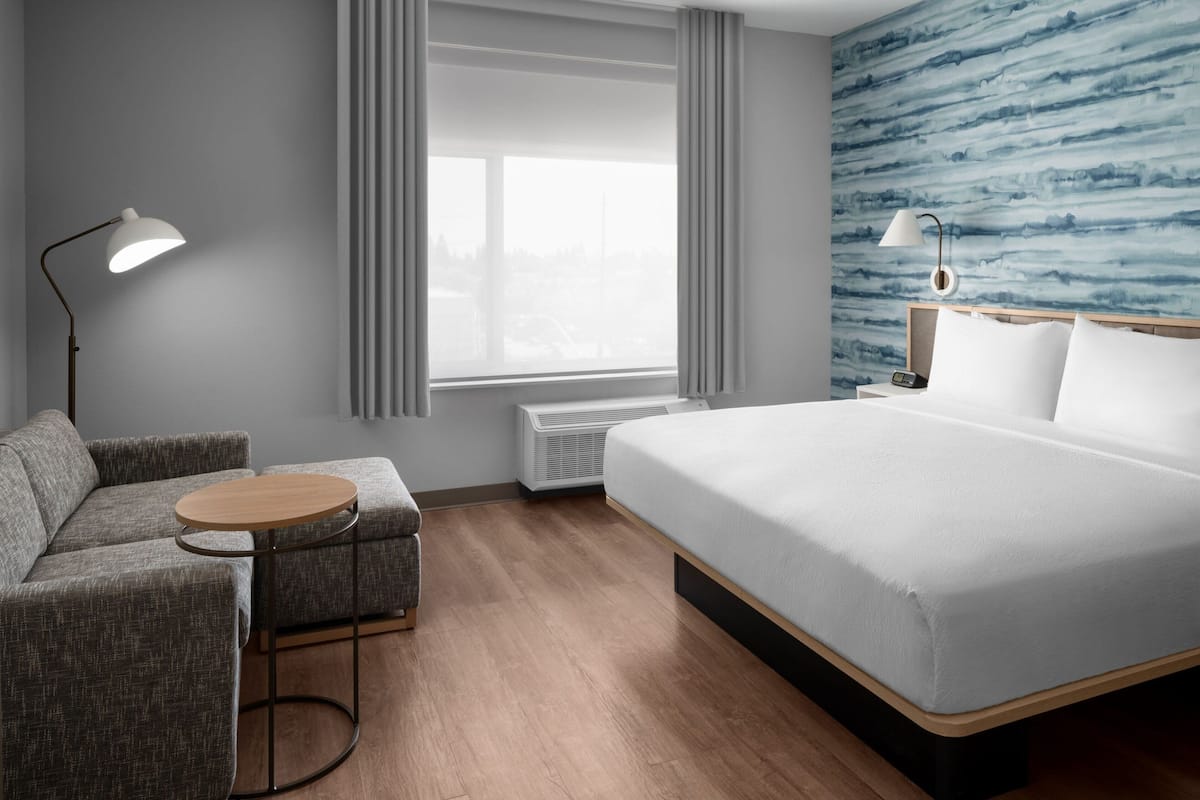 Een moderne hotelkamer met een bed, een bank, een klein rond tafeltje, een lamp en uitzicht op de stad door het raam.