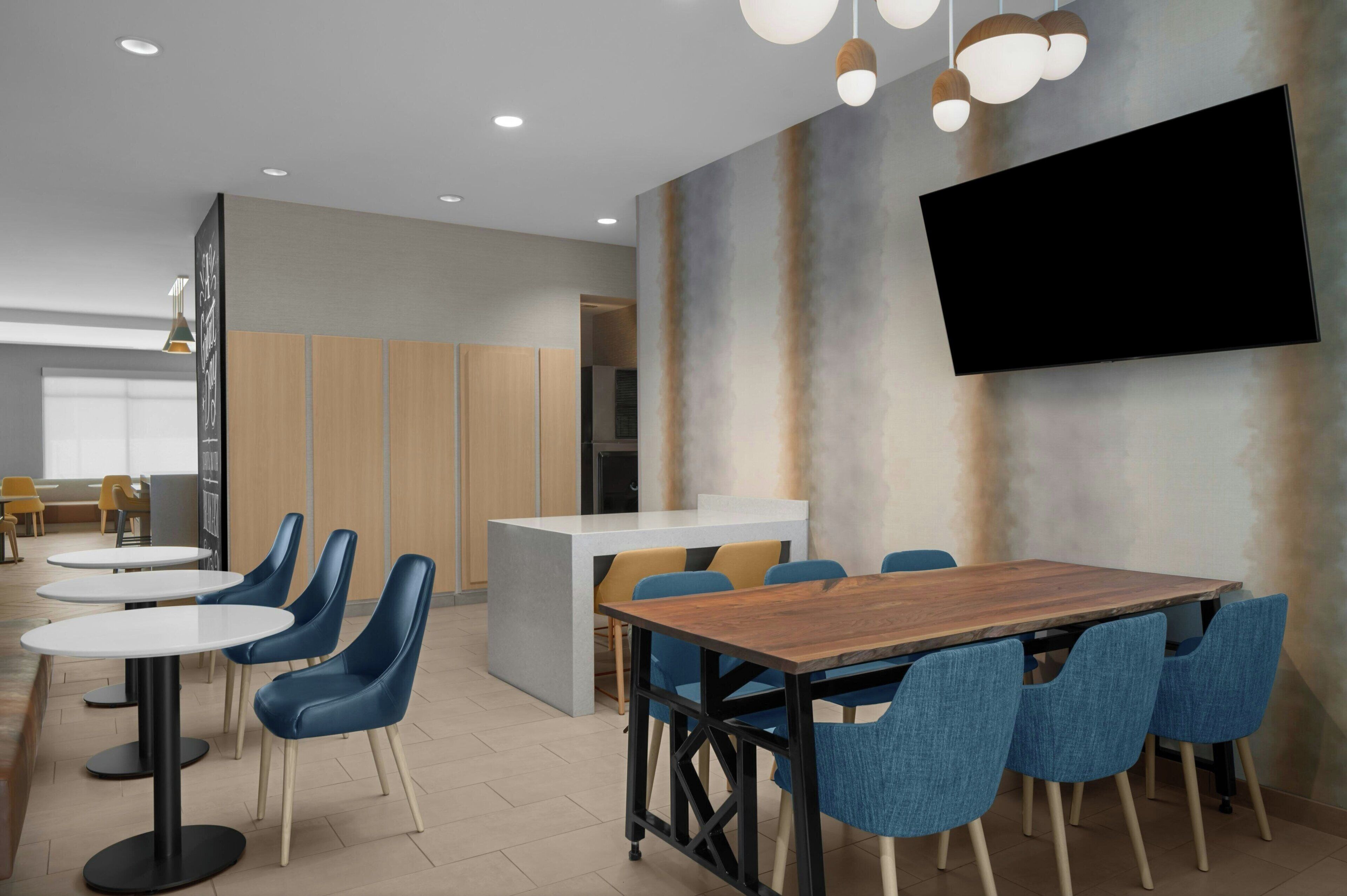 A modern dining area with round tables, blue chairs, a wooden dining table, and a flat-screen TV mounted on the wall.