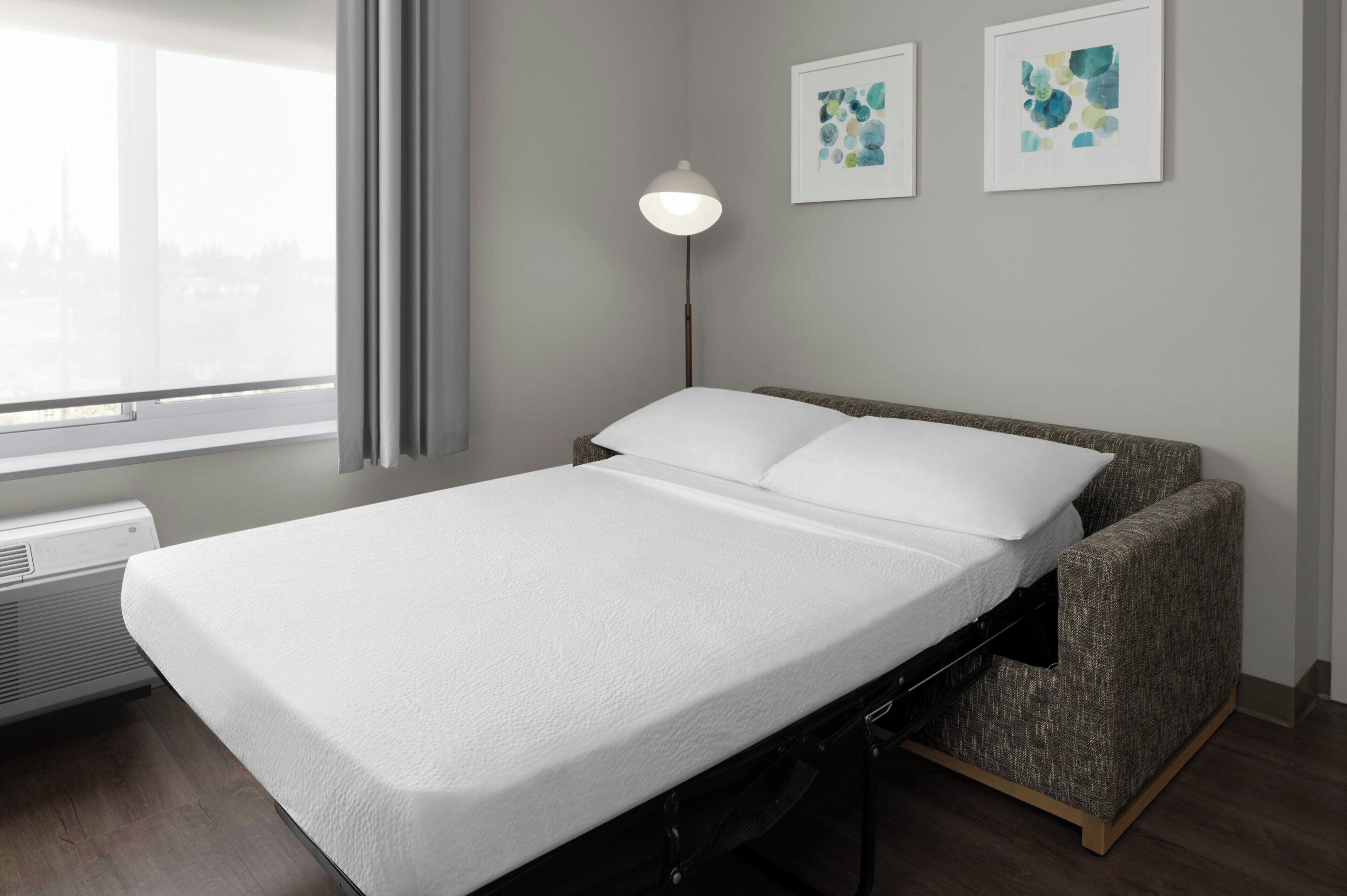 Una cama individual con colchón y almohadas blancos, una lámpara de noche y dos cuadros en la pared.