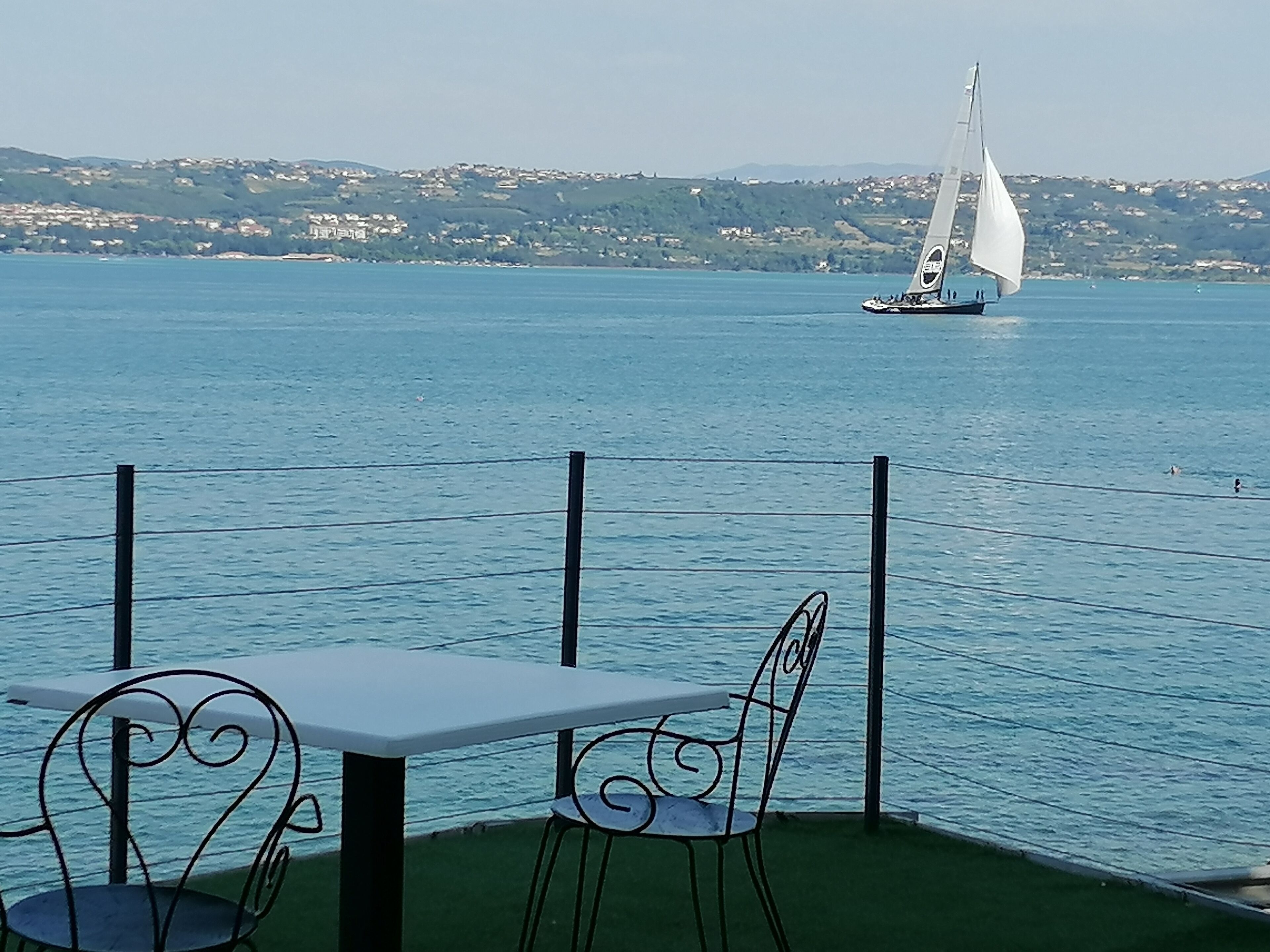 A table and chairs by a waterfront with a sailboat in the distance.