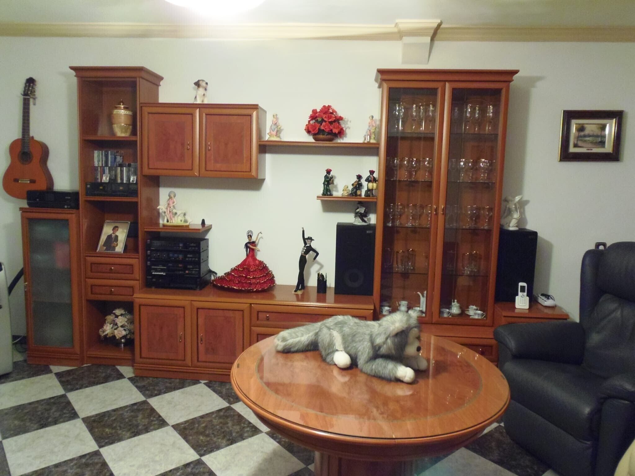 A living room with a checkered floor, a glass-top coffee table, a black leather sofa, wooden cabinets, a guitar, and various decorative items.