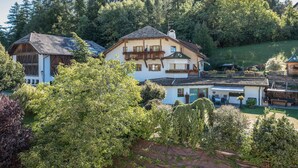 A traditional chalet with a balcony, surrounded by lush greenery and a forested hillside.