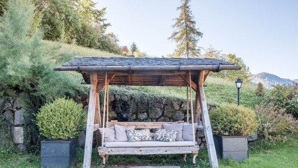 A wooden swing chair with cushions, surrounded by greenery and a stone wall.