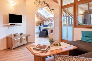 A cozy living room with a TV, wooden furniture, a dining area, and a view through an archway to a kitchen. - 'Lotschenhof Apartment Margerite' with Mountain View, Garden & Wi-Fi (Trodena)