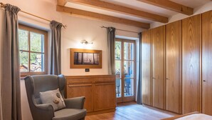 Une pièce avec un plancher en bois, un fauteuil gris, un lit, des meubles en bois et une fenêtre avec des rideaux.