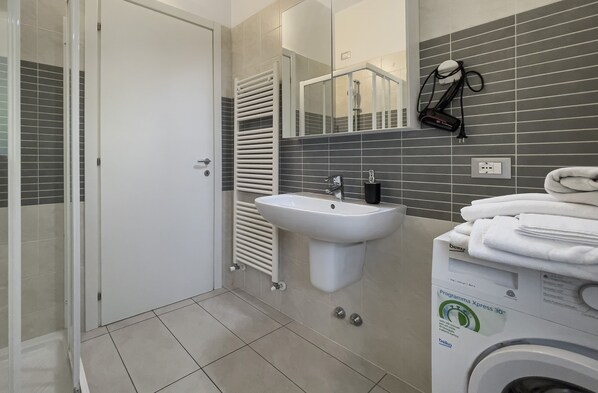 A modern bathroom with a white sink, a glass shower enclosure, a towel rack, and a washing machine.
