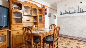 A dining area with a glass-top table, wooden chairs with cushioned seats, a wooden cabinet with glass doors, and a wall-mounted fire extinguisher.