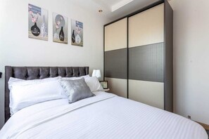 A bedroom with a tufted headboard, white bedding, and three wall-mounted artworks depicting vases with flowers. - Stay.Plus Splash of Glam Lavington (Nairobi)