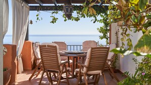 Eine Terrass mit Tisch und Stühlen, Blick auf das Meer und eine Pergola mit hängenden Pflanzen.