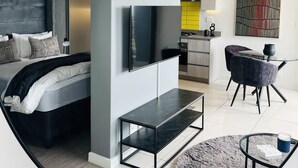 Un dormitorio moderno con cama, mesitas de noche, televisor y vista a la cocina.