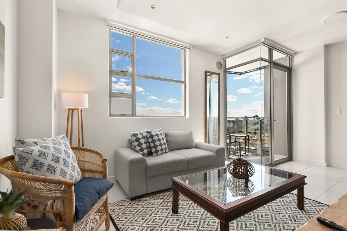 A modern living room with a grey sofa, a wicker chair, a glass coffee table, and a large window offering a city view.