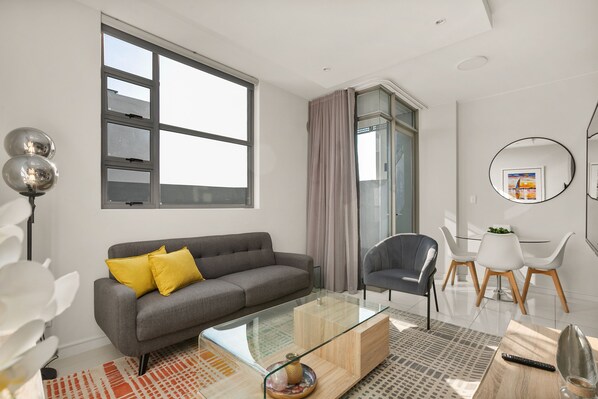 Un salon moderne avec un canapé gris, une table basse en verre et un coin repas comprenant des chaises blanches et une table en bois.