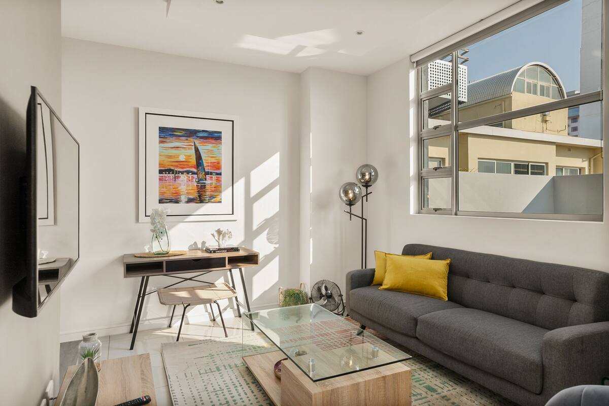 A modern living room with a glass coffee table, a grey sofa with a yellow pillow, a desk with a chair, a framed painting, and a large window with a view of buildings.