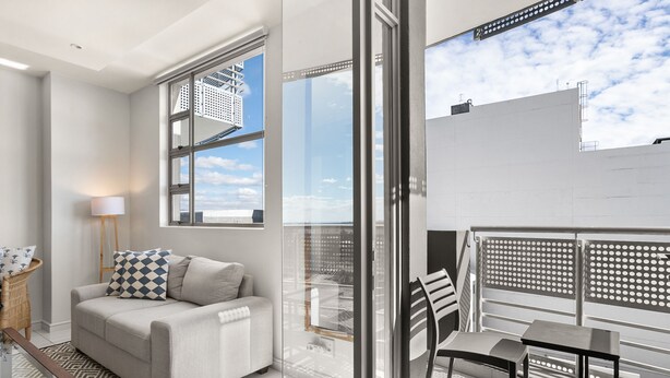 Una sala de estar moderna con un sofá, un ventanal de vidrio que da a un balcón y vistas del cielo y edificios.
