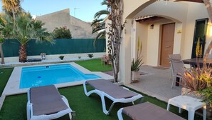 Un espace piscine aménagé avec des chaises longues, une petite table et une porte donnant sur un patio couvert.