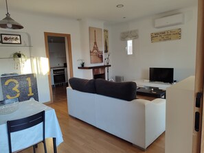 A modern living room with a white sofa, a dining table with a chair, a TV, and a fireplace.