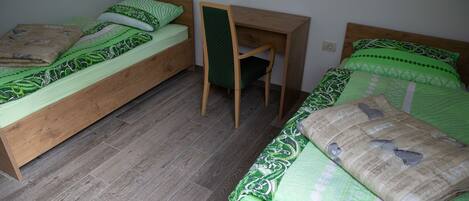 A small, compact room with two single beds, a wooden desk, and a chair.