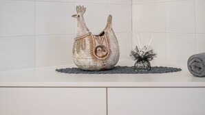 Une salle de bain avec un comptoir blanc, un coq décoratif en céramique, un vase en verre contenant une plante et une serviette enroulée.