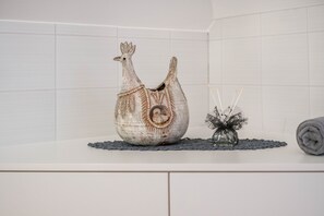 Une salle de bain avec un comptoir blanc, un coq décoratif en céramique, un vase en verre contenant une plante et une serviette enroulée.