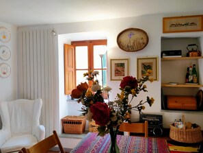 Een kamer met een witte stoel, een tafel met een kleurrijke tafelkleed, een vaas met bloemen, een rieten mand en een boekenkast met diverse spullen.