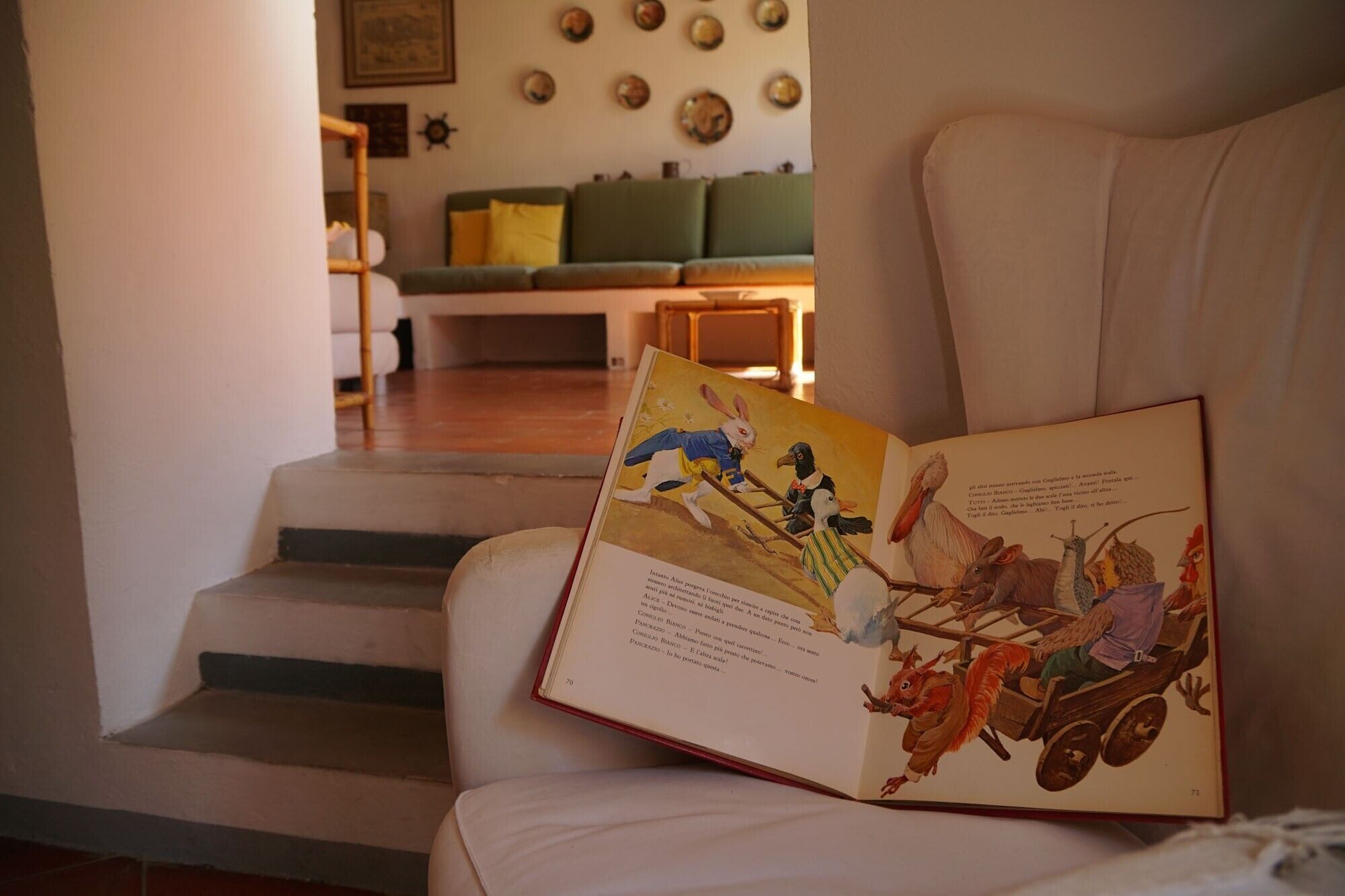 A book open on a chair in a living room with a staircase leading to another room.