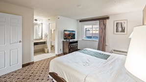 A hotel room with a large bed, a television, a bathroom with a mirror, and a window with curtains. - Coratel Suites 1 King Suite Oversized with Sofa Non-Smoking (Wichita)
