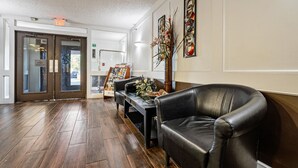 A lobby with a wooden floor, a black leather chair, a small table with a vase of flowers, and a wall-mounted artwork. - Coratel Suites 1 King Suite Oversized with Sofa Non-Smoking (Wichita)