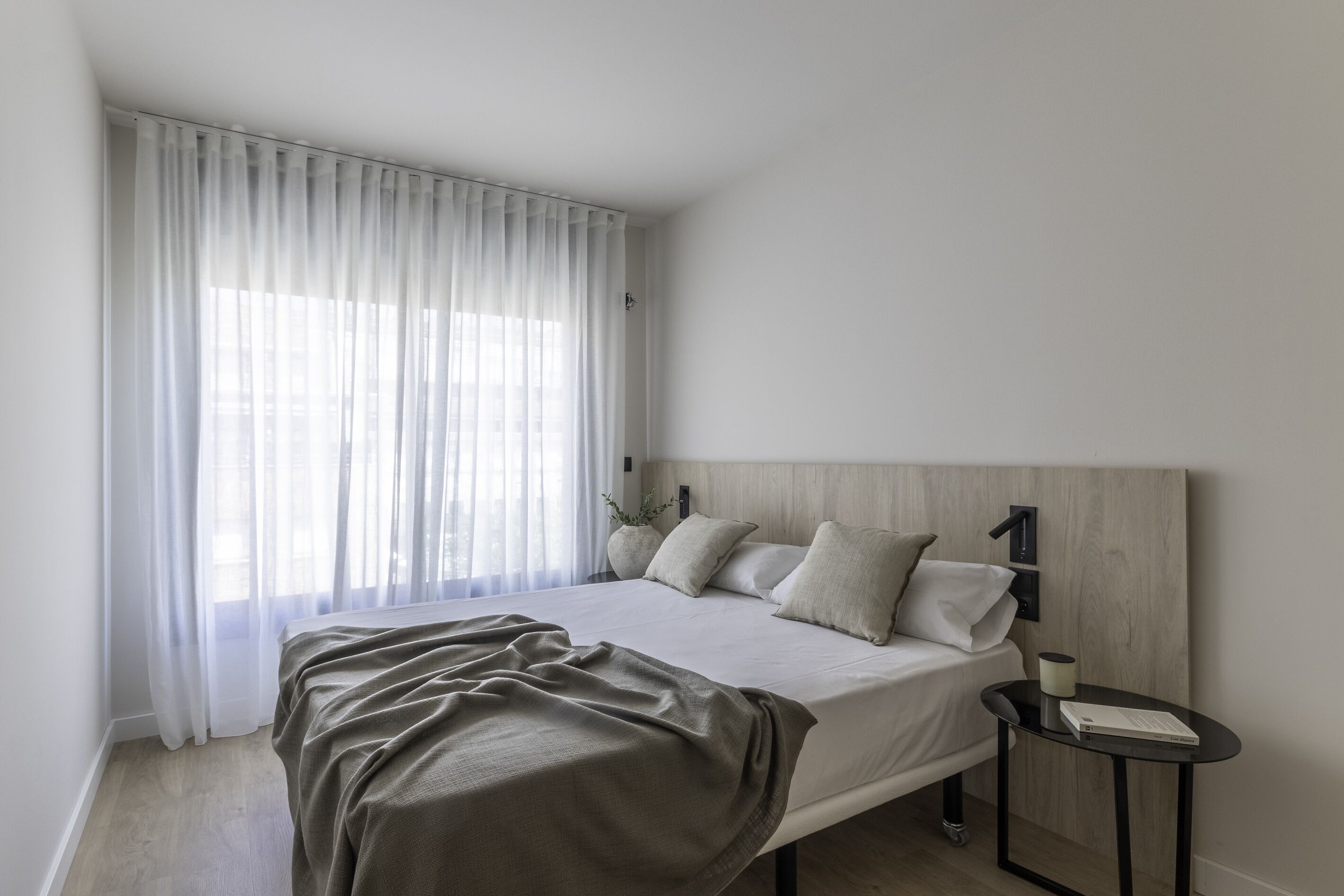A modern bedroom with a bed, a bedside table, a lamp, and a window with sheer curtains.