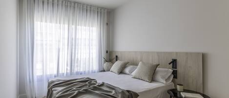 A modern bedroom with a bed, a bedside table, a lamp, and a window with sheer curtains.