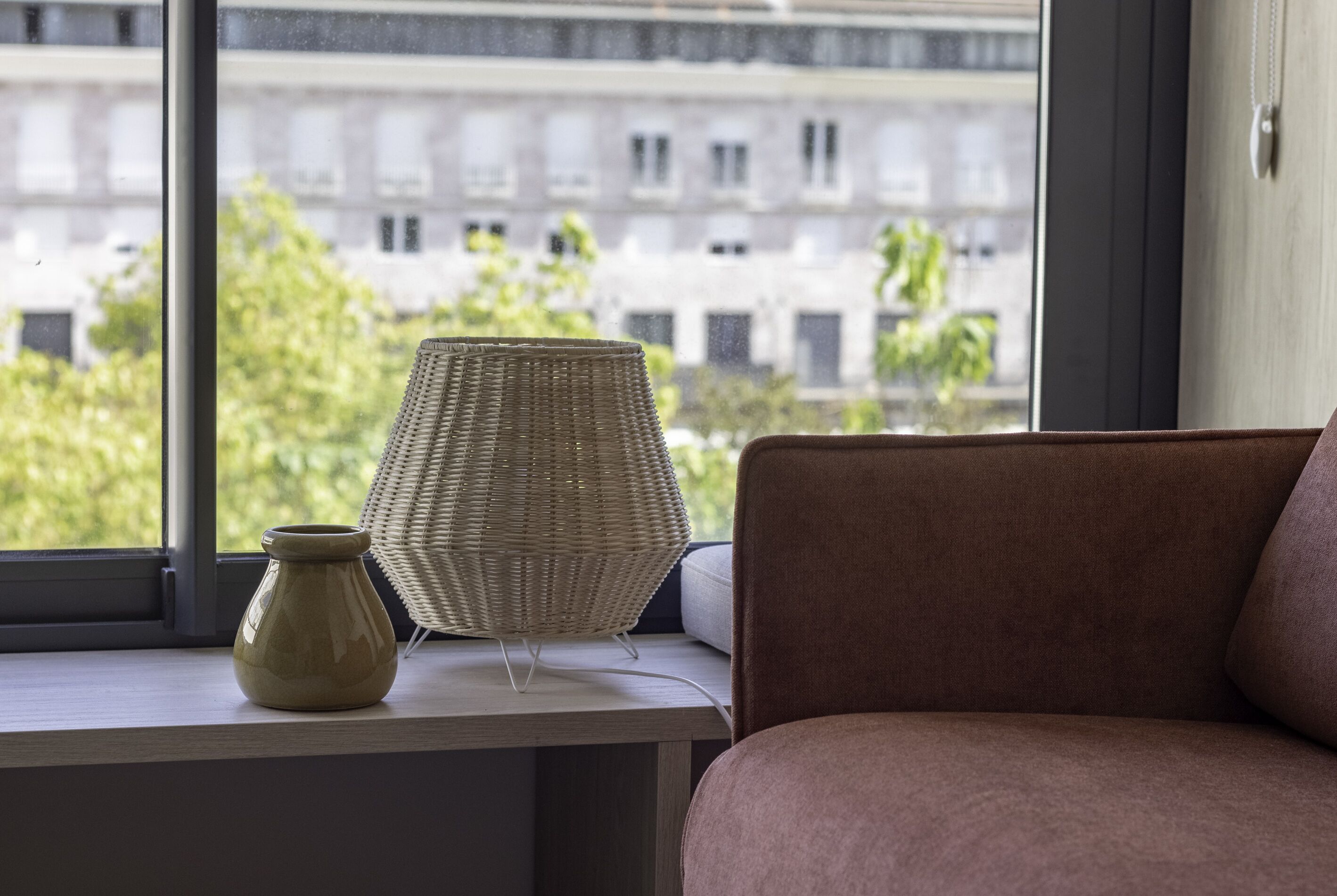 A room with a view of buildings through a window, a wicker lamp, and a vase on a table.