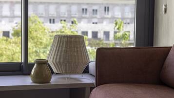 A room with a view of buildings through a window, a wicker lamp, and a vase on a table.