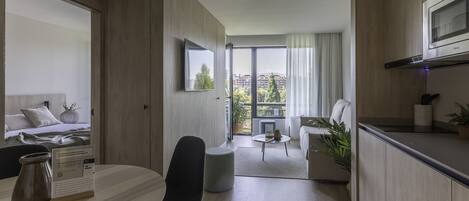 A modern hotel room with a bed, a dining table, a kitchen area, and a view of the cityscape.