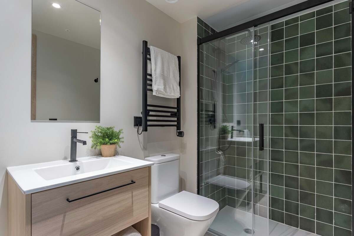 Un baño con un lavabo blanco, un perchero negro, un inodoro blanco y una cabina de ducha con mampara de cristal.