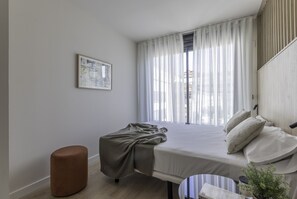 A modern hotel room with a bed, a red ottoman, a small table with a plant, and a window with sheer curtains. - Líbere Pamplona Yamaguchi (Pamplona)