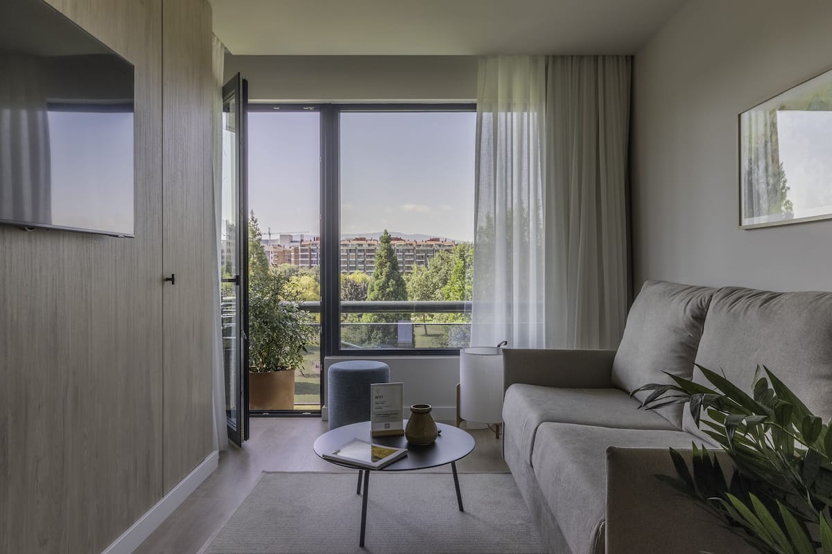 A modern living room with a sofa, a coffee table with a book and a vase, a potted plant, and a view of buildings through large windows.