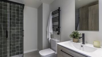 Un baño moderno con cabina de ducha de vidrio, inodoro blanco, toallero, lavamanos con espejo y una planta en maceta.