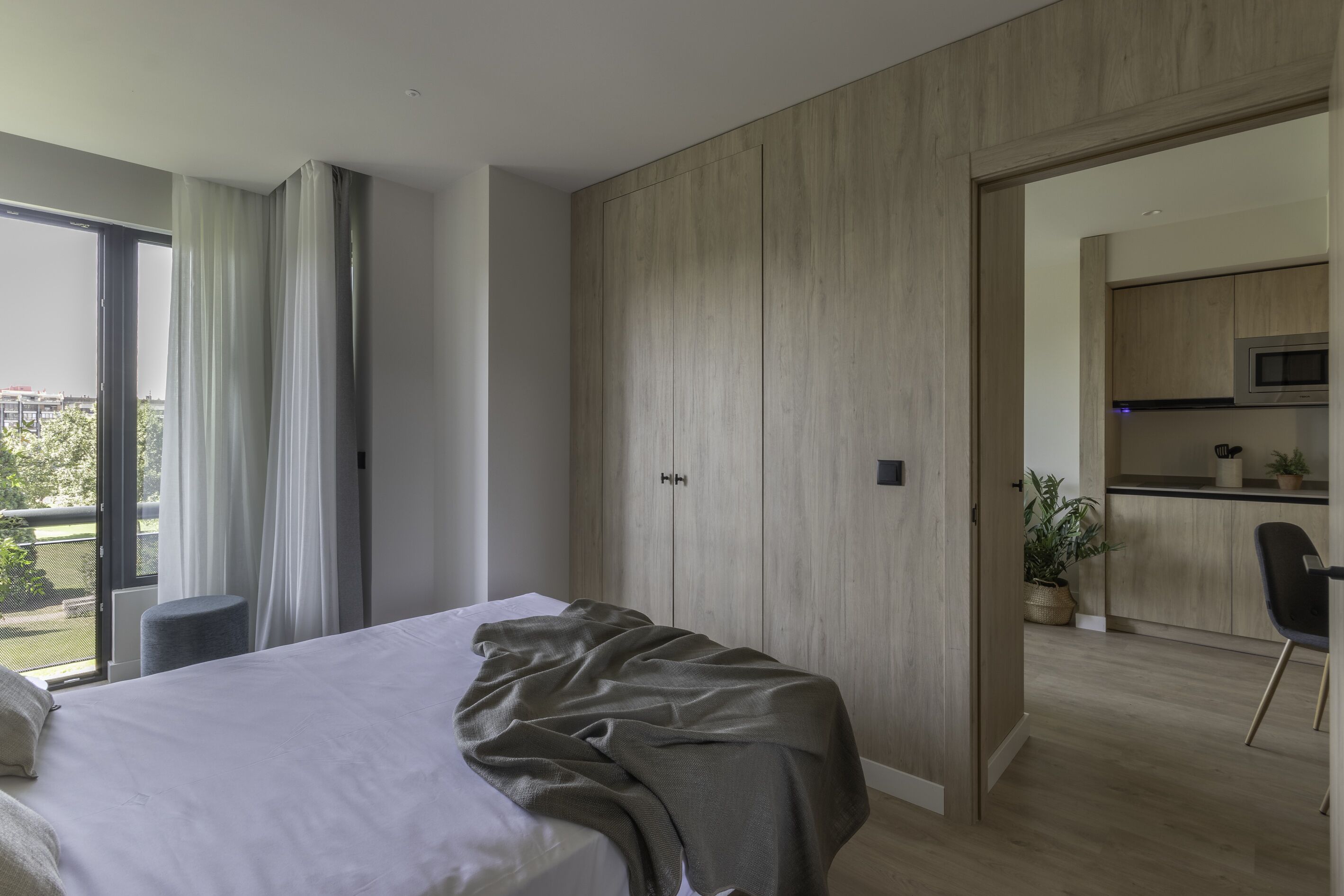 Un dormitori amb llit, vista a l'exterior, petita cuina i porta corredissa a una altra habitació.