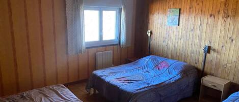 A room with wooden walls, a window with curtains, a bed with a blue bedspread, a bedside table, and a radiator.