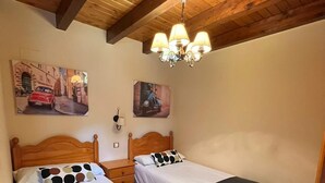 A room with two beds, wooden ceiling beams, a chandelier, and two wall-mounted paintings.