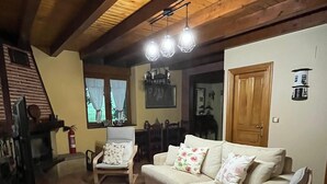 A cozy living room with a wooden ceiling, a beige sofa, a patterned armchair, and a dining area with a wooden table and chairs.