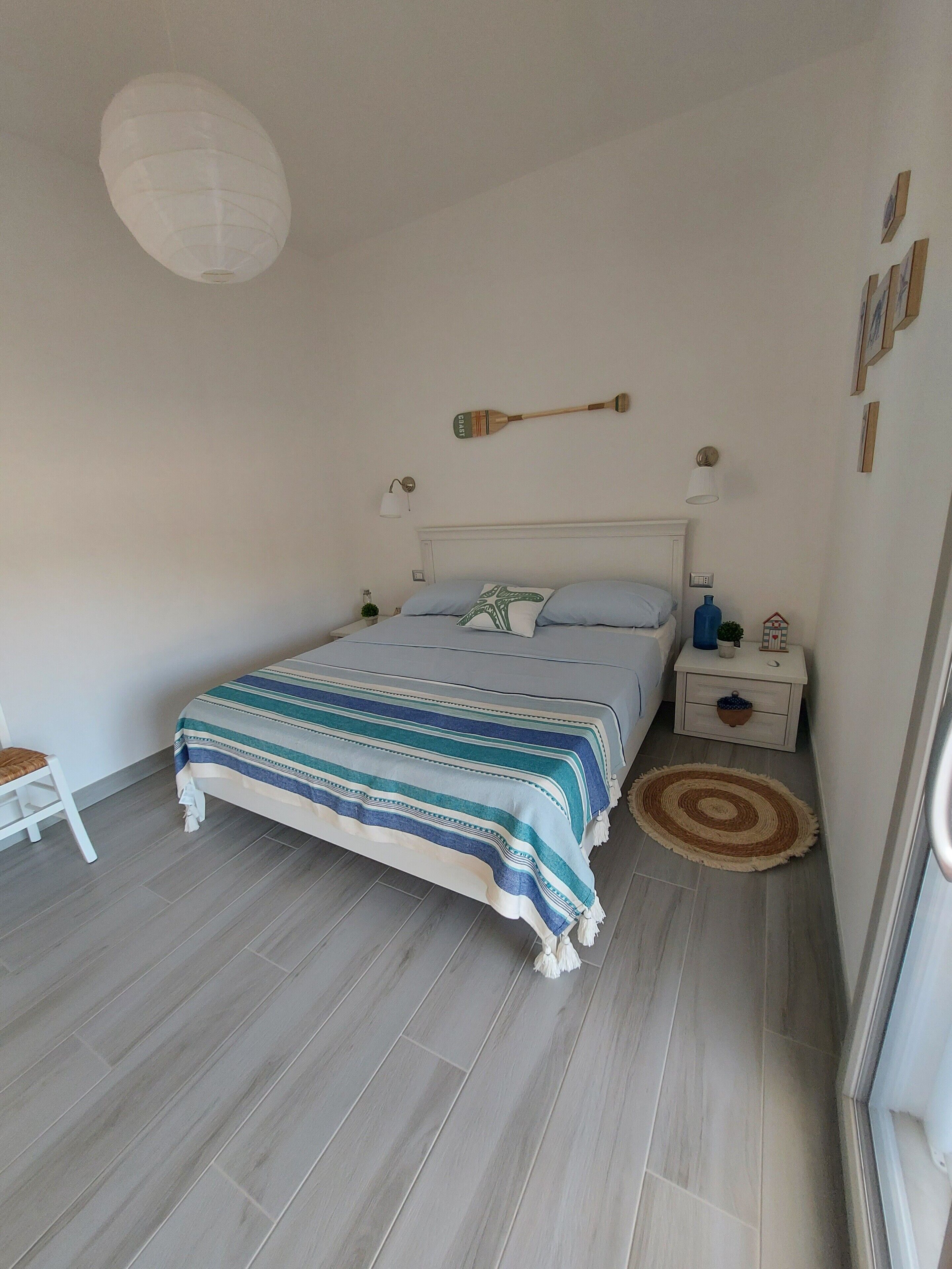 A bedroom with a bed, a nightstand, a lamp, a wall-mounted paddle, and a window.