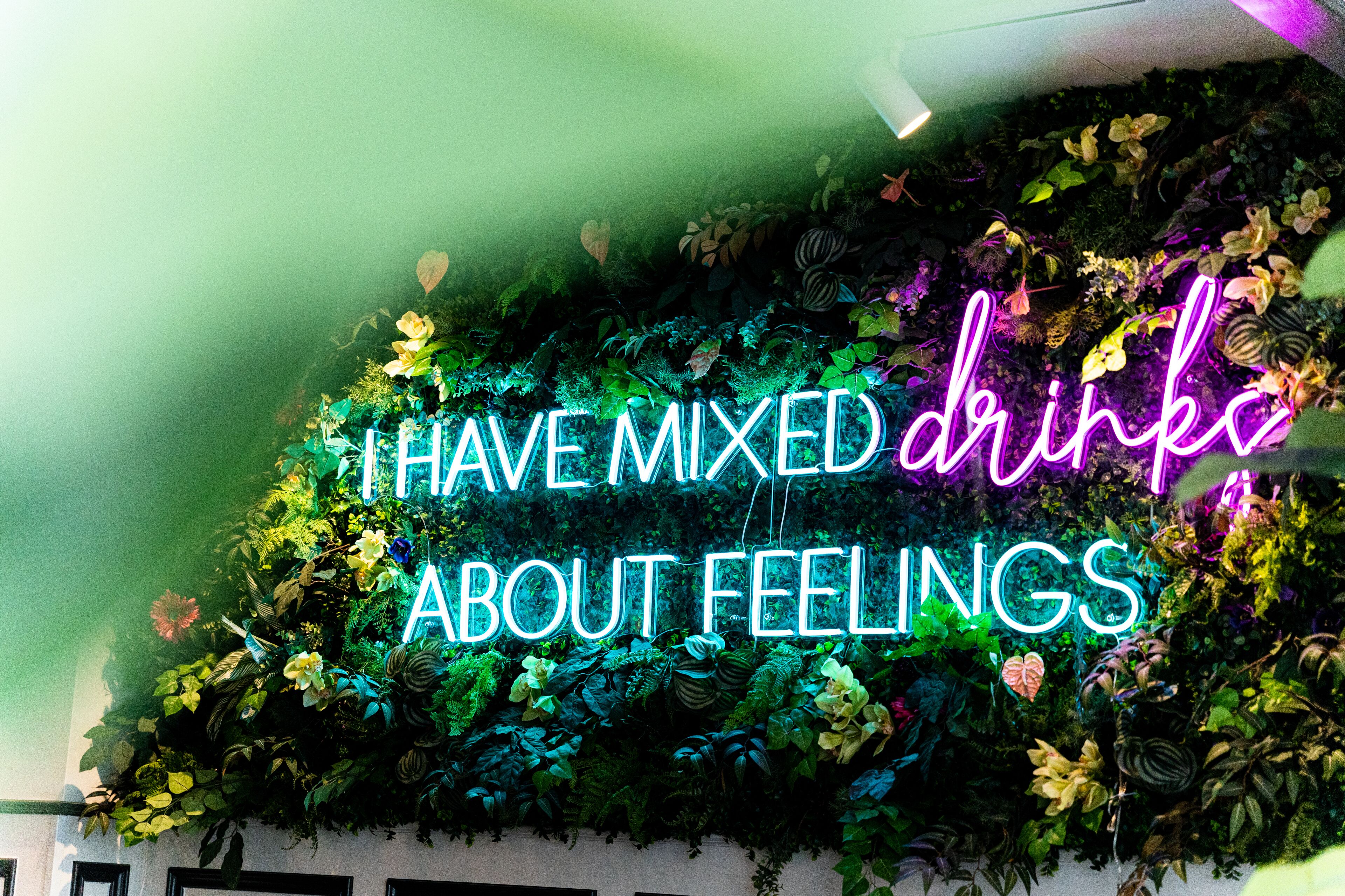 A neon sign with the text "I HAVE MIXED DRINKS ABOUT FEELINGS" against a backdrop of lush green foliage.