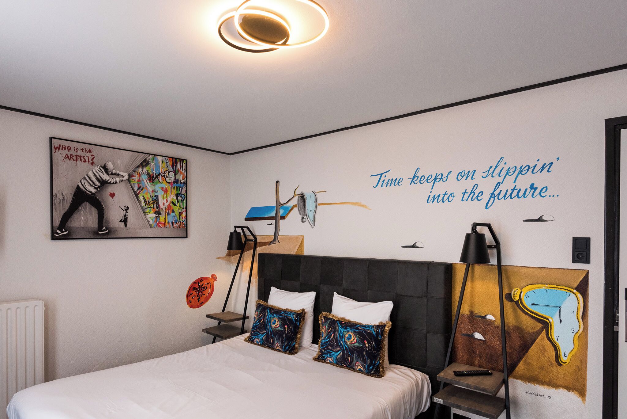 A modern bedroom with a bed, pillows, a nightstand, a lamp, and a wall with a clock and graffiti art.
