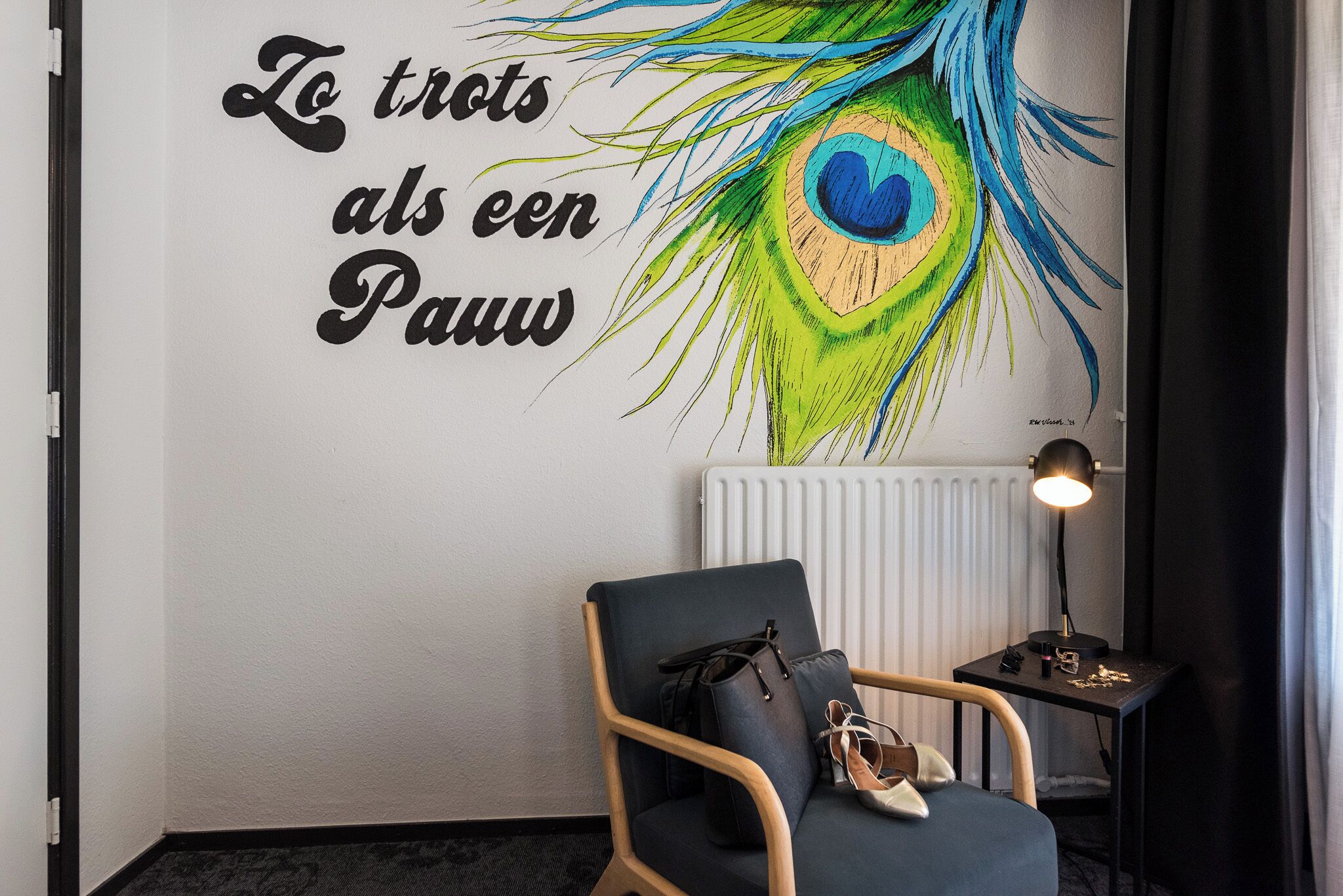 Une pièce dans laquelle on trouve une œuvre d ’art représentant des plumes de paon accrochée au mur, une chaise avec un sac à main posé dessus, et une petite table avec une lampe.